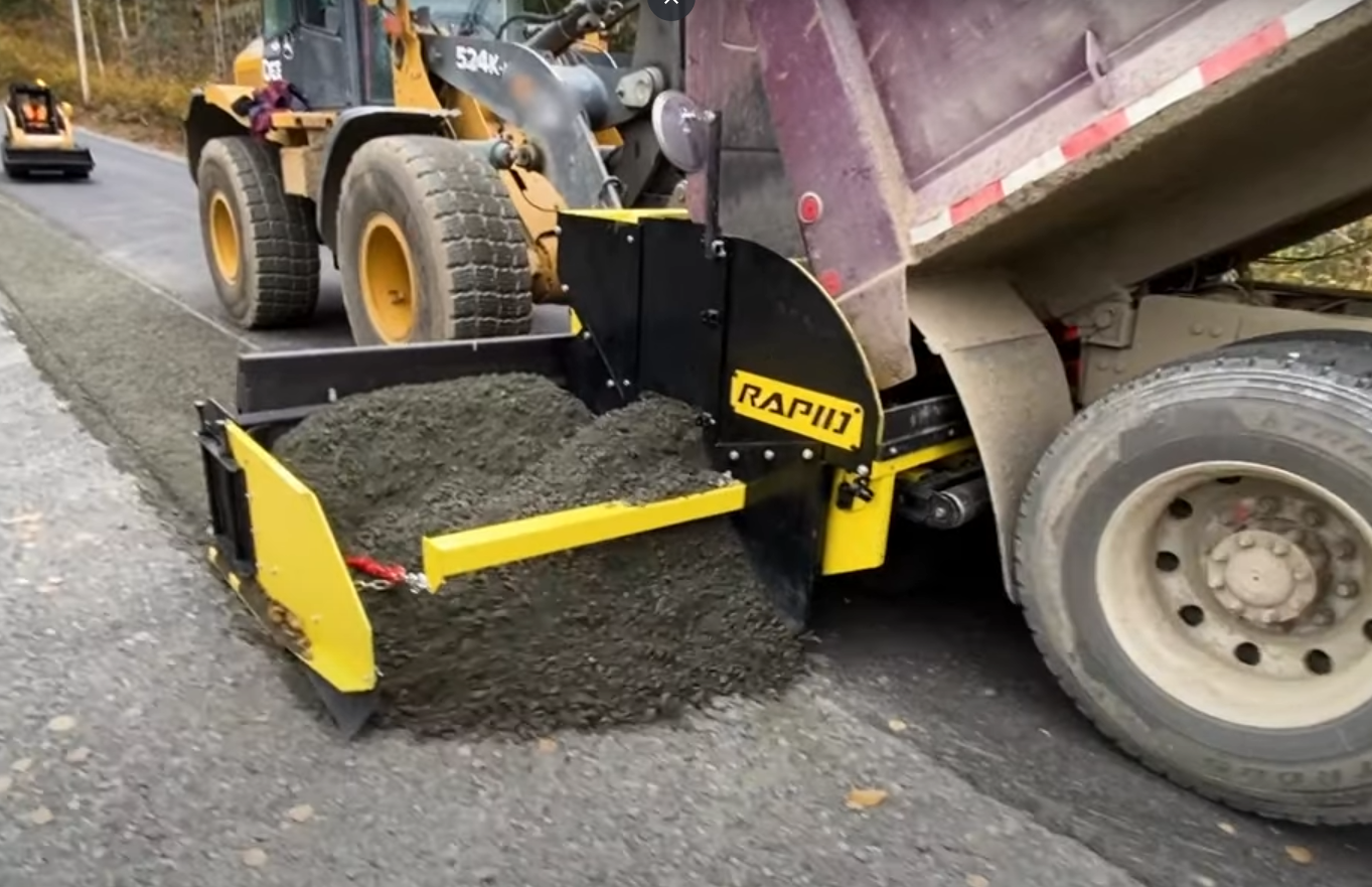 Chargeur qui effectue le pavage d'un accotement de route avec chargement par camion-benne.